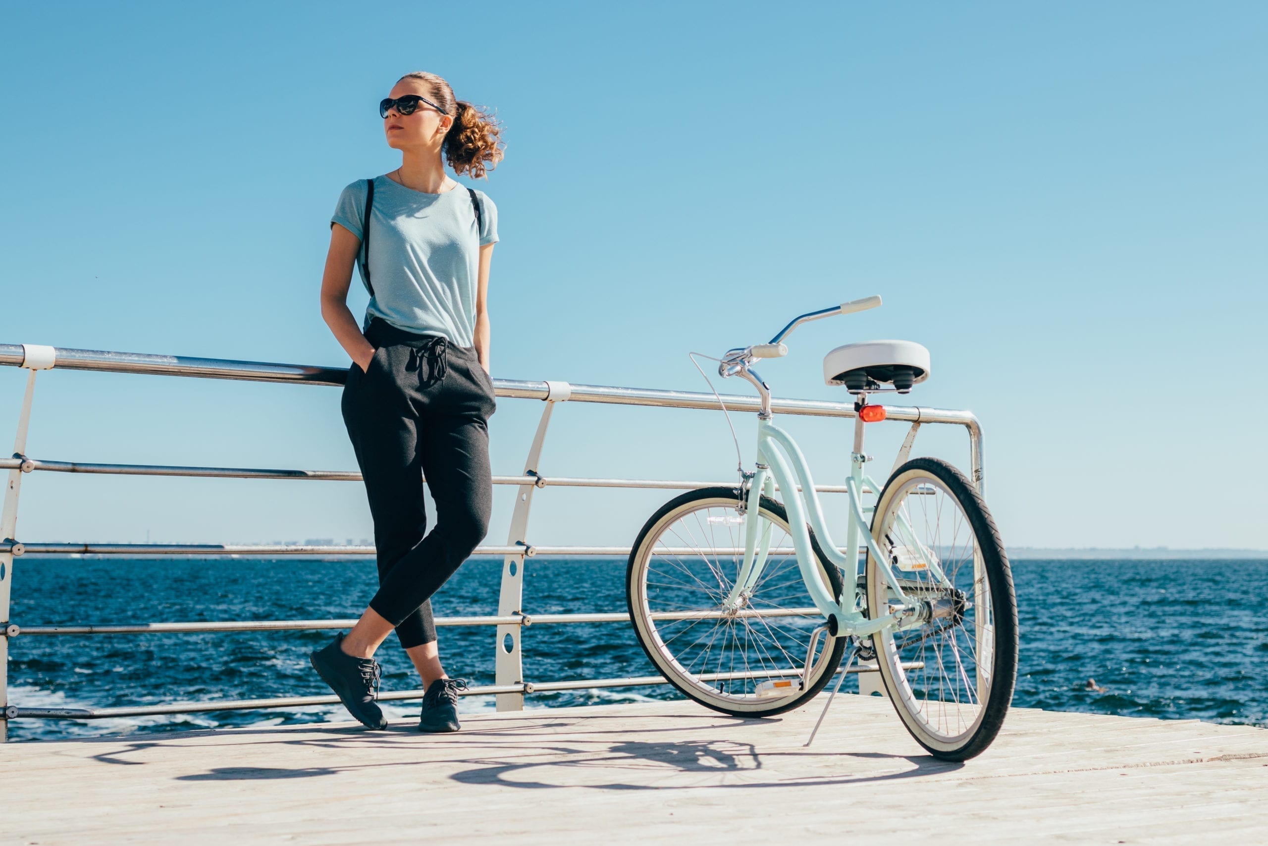 Full length image of young woman standing at background of blue sea near shiny cruiser bicycle. Female wearing casual outfit with hands in pockets resting after bike ride at sunny summer day.